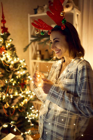 Cute joyful girl holding x-mas lights and dreaming about miracle on Christmas and New Year, woman wearing reindeer antlers headband at festive decorated homeの写真素材