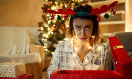Close up funny frowns face of festive x-mas woman. Disappointed woman with opened bad Christmas gift box at homeの写真素材
