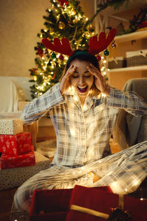 Surprised festive woman receive large present on Christmas time, looking amazed and happy, wearing reindeer antlers headbandの写真素材
