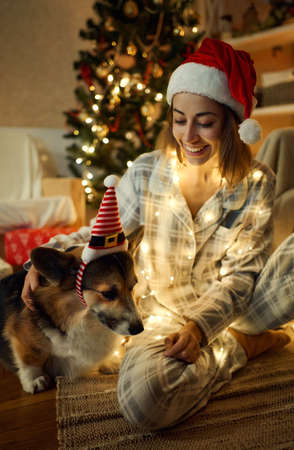 Happy woman in Santa Claus headwear with her cute pet Welsh Corgi dog sitting against decorated Christmas tree, lights and presents at homeの写真素材