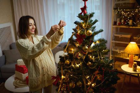 Pretty Woman in cozy winter sweater decorating christmas tree at home, holds christmas toy golden ball to decorate prepares for New Year timeの写真素材