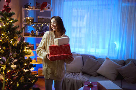 Happy smiling woman holds christmas gift boxes by decorated christmas tree on blue highlights background at festive home interiorの写真素材