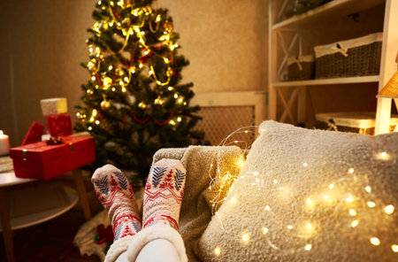First person view of woman legs in xmas socks lying on couch at cozy decorated home interior with...