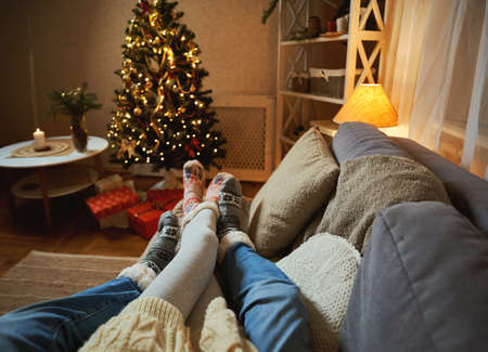 Winter holidays. Couple resting at home decorated with lights and Christmas tree, photo of feet in woolen xmas socksの写真素材