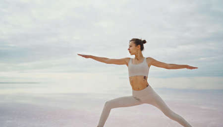 Side portrait confident athletic woman doing yoga exercise at early morning with beautiful natural calm background. Reflection cloudy sky in pink lake waterの写真素材