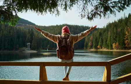 Back view backpacker woman sits on pier with open arms by mountain lake, demonstrating delight and freedom. Traveling to wild nature beautyの写真素材