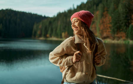 Cheerful traveler woman with backpack looking away and smiling, hiking near beautiful lake with forest on backgroundの写真素材