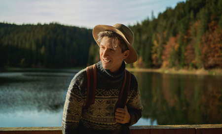 Portrait joyful traveler male in hat looking away and smiling, hiking near beautiful lake with forest on backgroundの写真素材