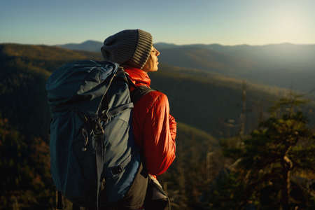 Back view woman hiker in bright red jacket with backpack standing on edge of cliff against background of sunset sky over mountainsの写真素材