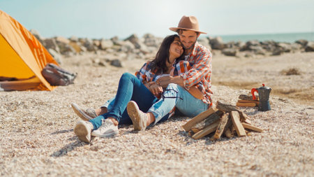 Happy traveling couple in love laughing, having conversation outdoors, sitting by firewood next to tentの写真素材