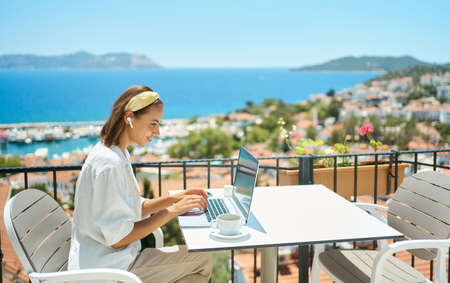 Young woman using laptop computer at cafe balcony of resort hotel with sea view, working typing emails browsing online enjoying drinking coffeeの写真素材