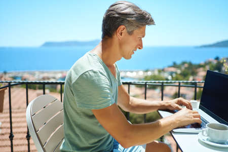 Portrait adult gray-haired man using laptop computer typing emails sending messages enjoying working from resort cafe with beautiful sea view.の写真素材