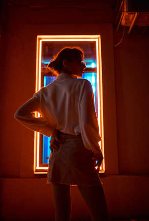 Cinematic urban night portrait of woman and neon light, fashion hipster girl posing against window frame with colored lights illuminationの写真素材