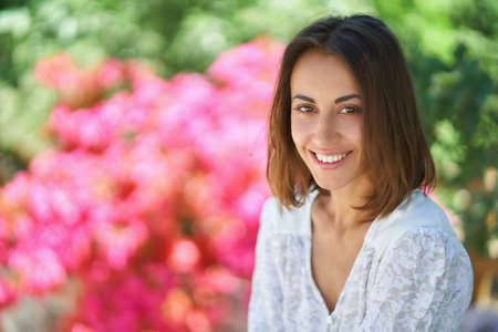 Beautiful woman with charming smile looks into camera. Close up outdoor portrait girl in spring with pink flowers and greens on backgroundの写真素材