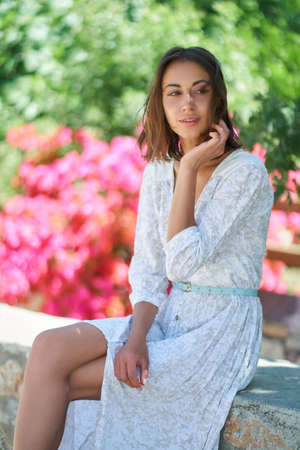 Attractive thoughtful young woman in white elegant dress sitting at garden with flowers. Spring is comingの写真素材