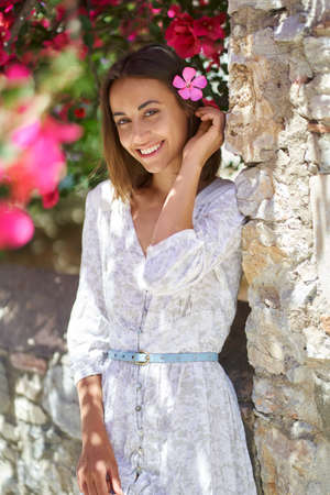 Smiling beautiful woman with flower in hair wearing white dress playful looking to camera outdoorsの写真素材