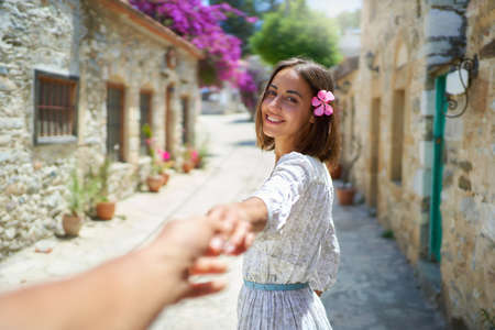 Adorable brunette woman with flower in hair smiling and holding hand of her boyfriend while walking at old city streetの写真素材