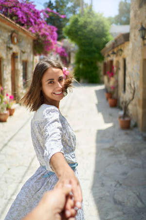 Smiling beautiful woman with flower in hair wearing white dress holding hand of her boyfriend, enjoying walking during vacationの写真素材