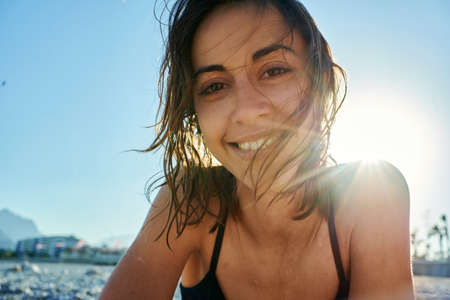 Young smiling woman at beach looking at camera. Close up portrait of beautiful girl with sunlight at seashoreの写真素材