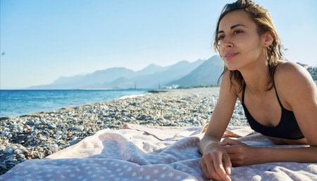 Beautiful thoughtful woman lying down on sea beach with mountains and looking away during summer...