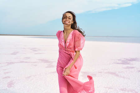 Beautiful happy woman emotionally smile to camera, wearing pink dress, enjoying time on desert beach of pink lake. Ukraine Sivash lakeの写真素材