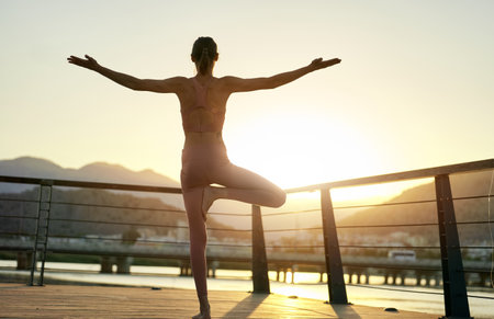 Back view silhouette athletic woman standing on bridge with open arms in Vrksasana Posture, Tree Pose. Practicing yoga during sunset with nature viewの写真素材