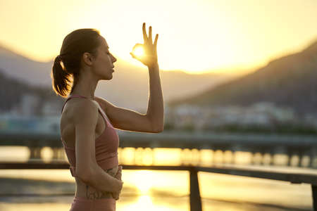 Young sporty woman practicing yoga and meditation outdoor during sunset against mountain range. Girl makes mudra gesture with sunlightの写真素材