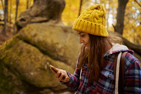 Woman hiker in checkered jacket and yellow beanie hat with backpack using smartphone at woods during active outdoor vacationの写真素材