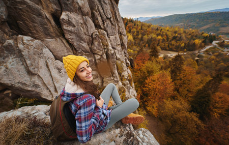 Traveler millennial girl in yellow beanie hat with backpack sitting on cliff edge with autumn forest and enjoying beautiful valley view. Adventure vacationsの写真素材