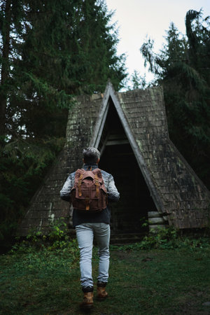 Rear view traveler man with backpack coming to old vintage wooden hut in dark woods. scandinavian style moody aesthetic imageの写真素材