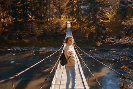 Fall season outdoor portrait woman walking on hanging bridge over river, girl turning around and giving hand to camera like follow meの写真素材