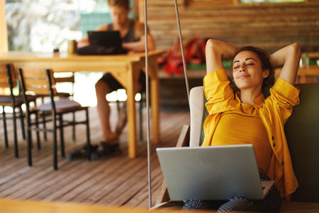 Carefree woman relaxing on swing in cafe shop or coworking hub with her laptop computer. Vacations and sometimes working remotelyの写真素材