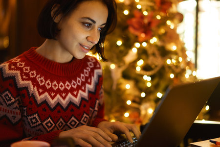 Caucasian female blogger in red winter sweater smiling indoors while texting and reading connected to 4g wireless in coffee shop xmass interiorの写真素材