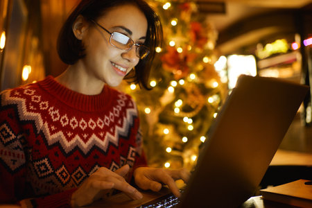 Beautiful smiling woman in eyeglasses with laptop sitting in cafe with christmas tree festive decoration. Working before holidays, work deadlineの写真素材