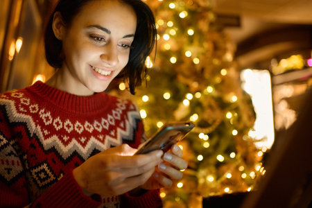 Closeup of pretty smiling woman using smartphone in cafe with christmas festive tree and lights. Girl watching video, chatting in social networkの写真素材