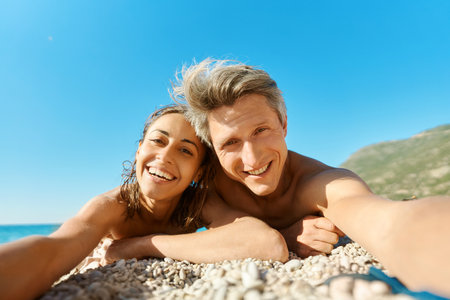 Portrait of joyful smiling couple lying on sunny beach, making selfie picture on romantic honeymoon holiday or vacation travelの写真素材