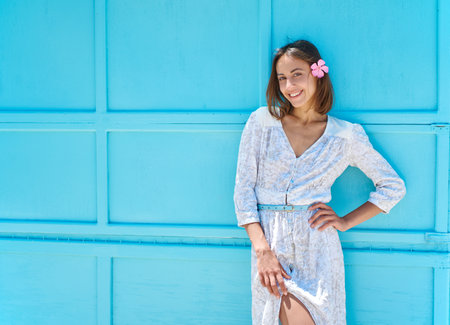 Charming young lady on bright blue background. Beautiful female model with flower in hair wearing white dress posing at camera and smiles broadly with teethの写真素材