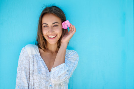 Close up face beautiful laughing girl on bright blue background. Femininity model with flower in hair posing smiling to cameraの写真素材