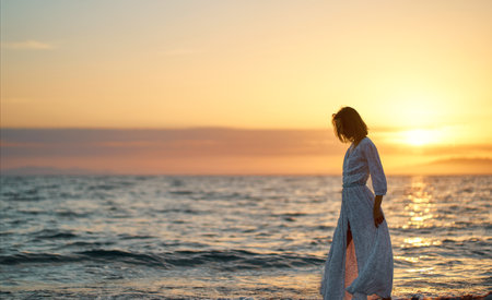 Beautiful young woman in long elegant dress at sunset. Silhouette of dreamy girl walking along the seaの写真素材