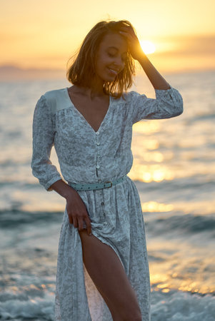 Beautiful elegant young woman in dress standing in front of sea at sunset. Nature and beauty conceptの写真素材
