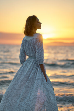 Gorgeous freedom female in romantic elegant white dress in front of serene sunset sky at seashoreの写真素材