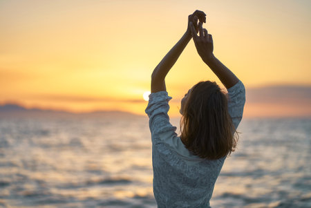 Back view carefree woman with hands up to the sky at sunset over the sea. Silhouette of dreamer girl looking hopeful at the horizonの写真素材