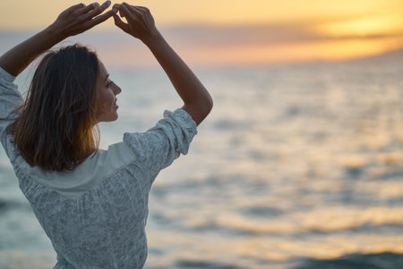 Happy young woman posing with hands up to the sky, enjoying amazing sunset over the seaの写真素材