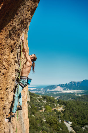 Vertical image athletic male climbing on the high rock with amazing mountains and blue sky behindの写真素材