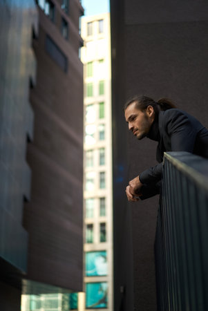 Pensive young man dressed in suit looking away while leaning on railing against a backdrop against tall dark buildings. The image evokes feelings of contemplation, introspection, and the pressures of modern urban life.の写真素材