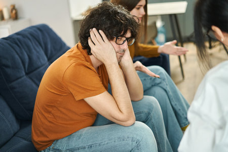 Couple having therapy session with a psychologist to solve their relationship problems and crisis. Sad man covering his ears with hands to avoid hearing abuseの写真素材