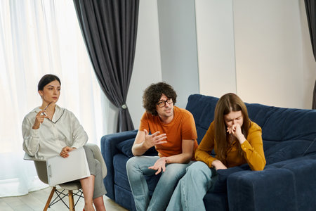 Emotional and dramatic moment in therapy session. Man expressive shouting at his wife, woman is overwhelmed with tears and despair. A skilled psychotherapist observing and trying understand and resolve conflictsの写真素材