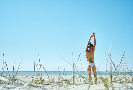 Escape to paradise. Feel the warmth of the sun. Bikini girl stands gracefully with her back turned, reaching her arms up towards the blue sky on white sandy seashoreの写真素材
