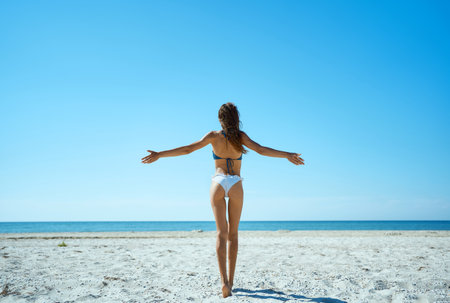 Tranquil beauty of secluded island getaway. Gorgeous sunkissed woman standing on the beach with open arms, blissful relaxation and harmony with natureの写真素材