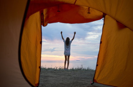Stunning morning landscape as seen from a tent, with glorious sunrise sky. A silhouette of woman joyfully raises her arms to welcome the dawnの写真素材